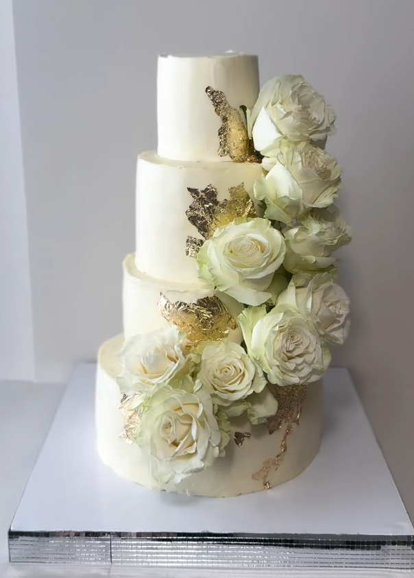 Four-tier white wedding cake with cream roses and gold leaf accents — handmade by Monali YYC